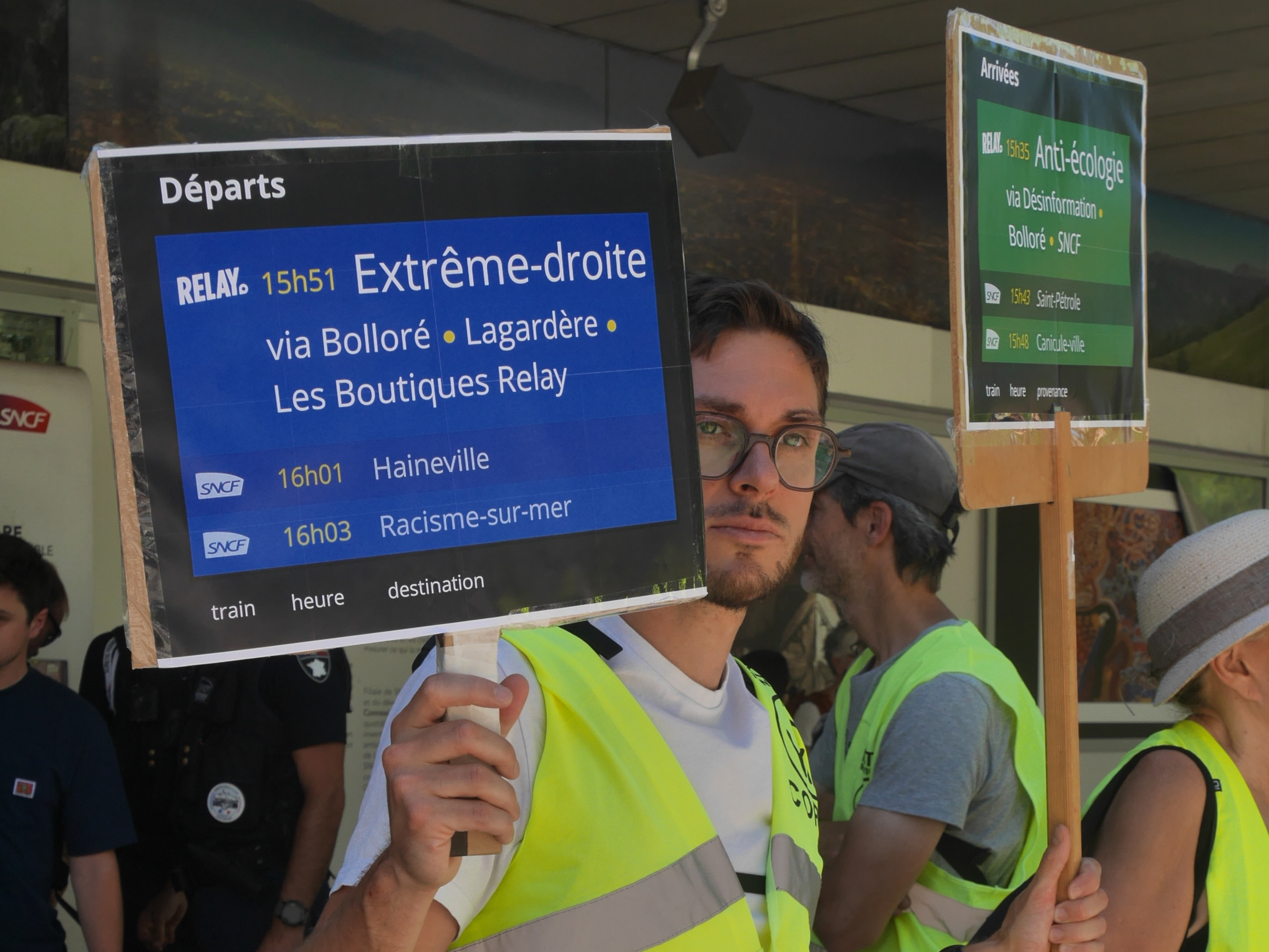 Un personne en brandit des panneau d'arrivée et de départ de trains détournée à la gare de Grenoble. 
On peut y lire des trains aux départ de "Extrême-droite via Bolloré - Lagardère - Les Boutiques Relay".